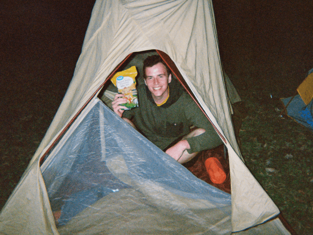 A film photo taken at night with flash of myself inside a tent, holding up a bag of trail mix.